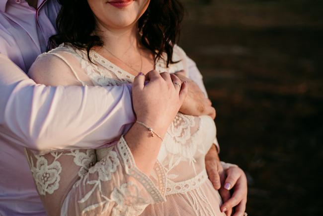 20180614-DSC_9440 A woman holding onto her partner's arm that is wrapped over her chest during a golden hour maternity photography session at The Pines Wanneroo in Perth