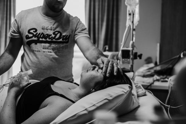 Black and white image of a man touching the forehead of his pregnant partner who is labouring in the hospital bed during a Perth birth photography session