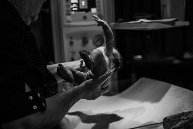 Black and white image of the midwife holding a new baby to be weighed during a Perth birth photography session