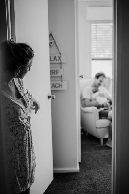 20180615-DSC_0233 Black and white image of woman peeking into the doorway of her husband holding her baby during lifestyle newborn photography Perth session