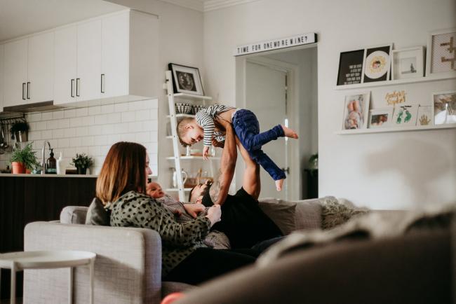 20180706-20180706-DSC_6070 Father lifting son in the air on the couch while mother holds new baby during lifestyle newborn photography Perth session