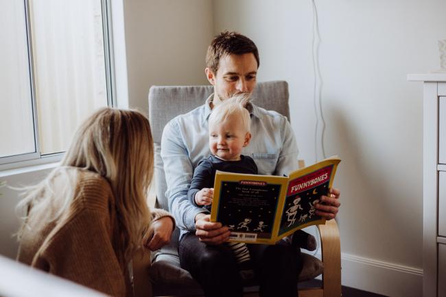 20180708-DSC_8005 little boy sitting on his dad's lap and smiling at his mother as his dad reads him a story during an in-home family photography Perth session