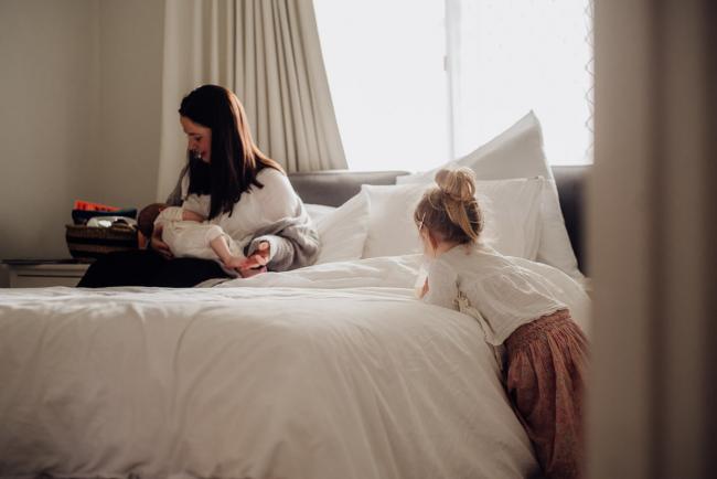 mother breastfeeding her new baby on the bed as her daughter leans against the bed during lifestyle newborn photography Perth session