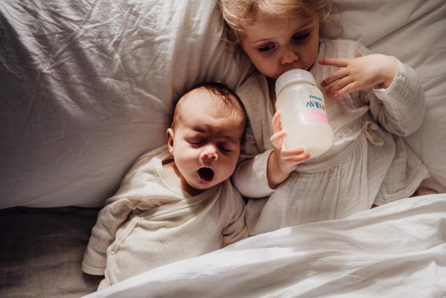 Baby yawning in bed next to her big sister who is drinking a bottle during lifestyle newborn photography Perth session