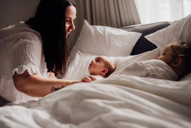 Baby and big sister laying in bed while mum leans over to them during lifestyle newborn photography Perth session