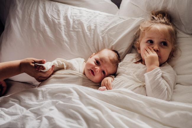 Baby and her big sister laying in bed during lifestyle newborn photography Perth session