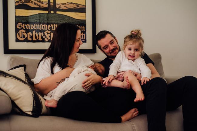 Mother breastfeeding her new baby while big sister sits on dad's lap during lifestyle newborn photography Perth session