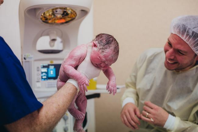 Midwife holding new baby up while dad smiles at her during Perth birth photography session