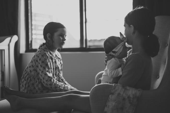 Black and white image of two siblings and their new baby sister - one sister is holding the baby on the feeding chair during a Perth newborn photography session