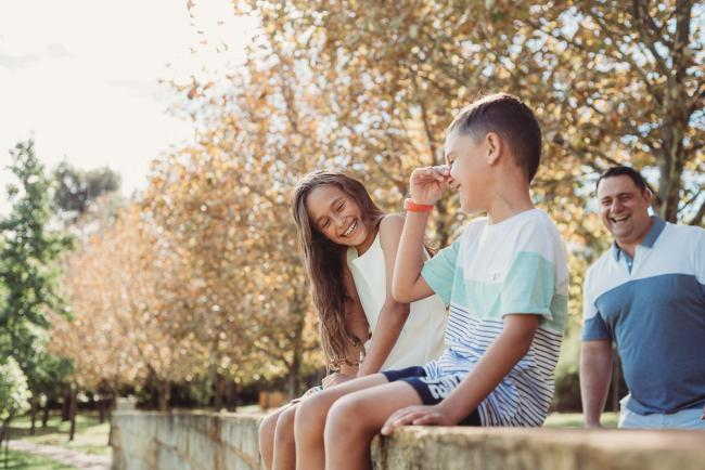Brother and sister laughing on a brick wall as their dad laughs behind them during a family photography session in Perth
