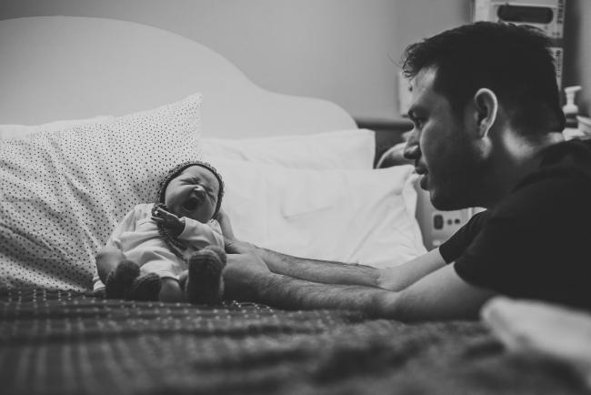Black and white image of dad propping baby up against pillows on the hospital bed during Perth Fresh 48 session