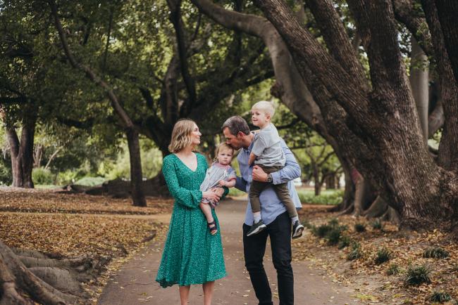 Mother holding her daughter and father holding his son as he leans over to kiss his daughter during a family photography session at Hyde Park in Perth