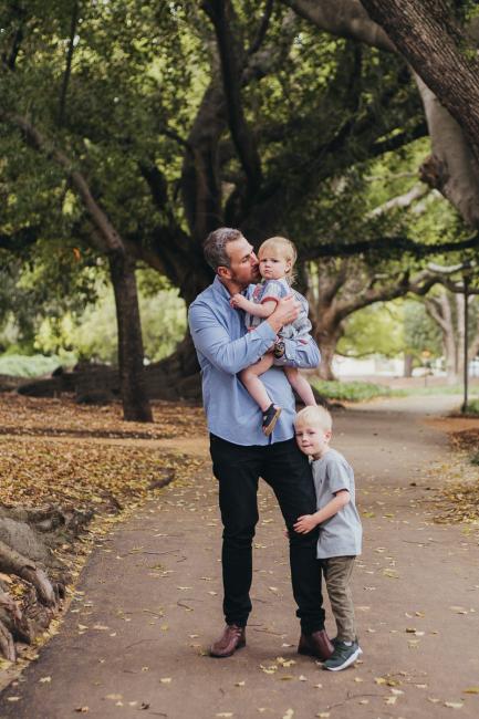 Dad holding his daughter whilst kissing her, and his son hugs his leg during a family photography session at Hyde Park in Perth