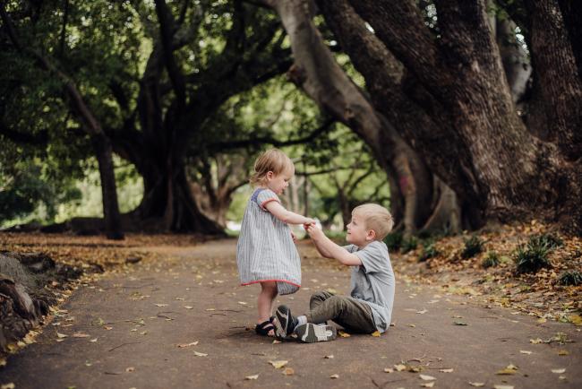 Brother sitting on the ground while his little sister tries to lift him up with her hands during a family photography session at Hyde Park in Perth