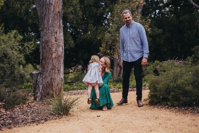 Dad standing, and mum kneeling on the ground as she lifts her daughter during a family photography session at Hyde Park in Perth