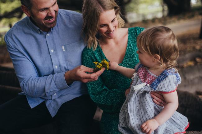 Mother and father smiling at their daughter as she hands them yellow flowers during a family photography session at Hyde Park in Perth