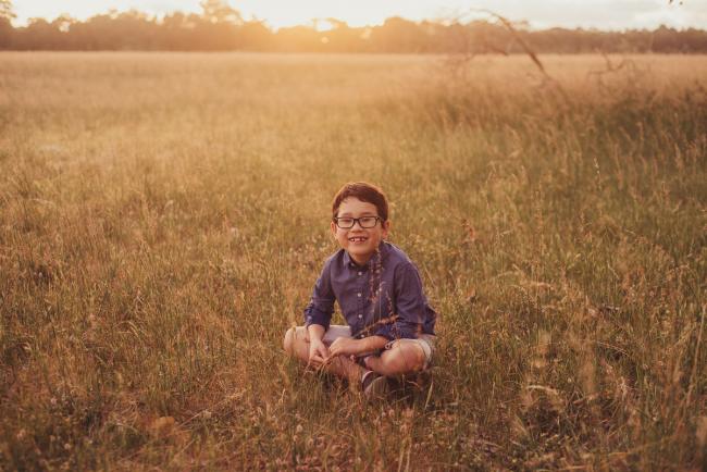 Little boy sitting cross legged and smiling in a field during a golden hour family photography session at Perry's Paddock in Perth