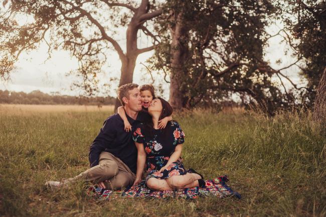 Mum and dad sitting on the grass with their daughter standing between them during a golden hour family photography session at Perry's Paddock in Perth