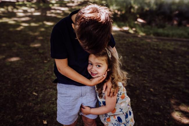 Big brother wraping his hands around his little sisters face during a mini session with Perth family photographer at Hyde Park