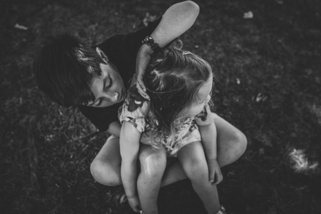 Black and white image of a little girl sitting on her big brothers lap while he plays with her hair during a mini session with Perth family photographer at Hyde Park