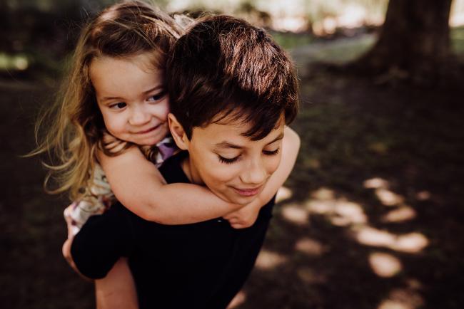 Little girl being piggy backed by her big brother during a mini session with Perth family photographer at Hyde Park