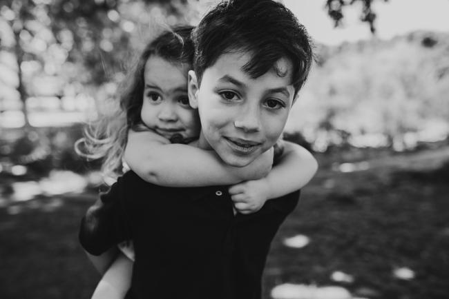 black and white image of a little girl being piggy backed by her big brother during a family photography session in Hyde Park, Perth