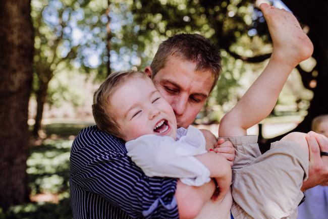 Little boy laughing and being held and tickled by his dad during a mini session with Perth family photographer at Hyde Park