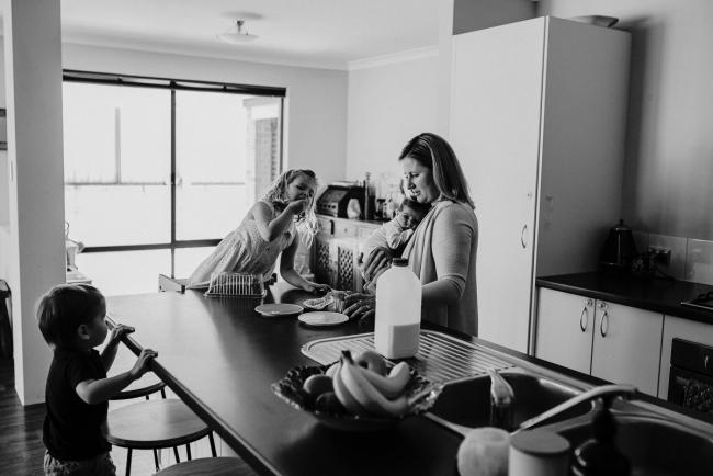20181111-DSC_8207-1 black and white image of mother multitasking in the kitchen with her new baby and her two children in a lifestyle family photography session Perth