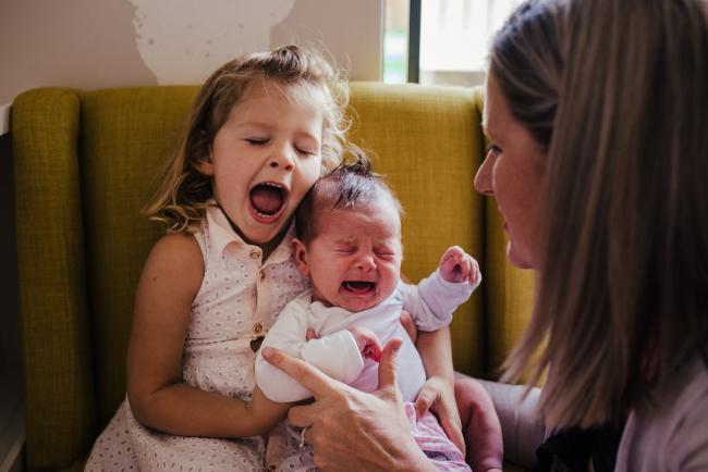 20181111-DSC_8235-2 Big sister pretending to cry as she holds her crying baby sister on a yellow chair in a lifestyle family photography session Perth