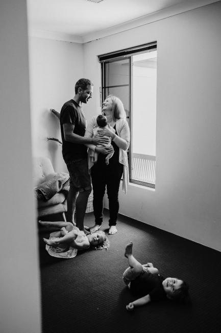 20181111-DSC_8304 black and white image of two parents standing with their new baby while their other two children roll on the floor in a lifestyle family photography session Perth