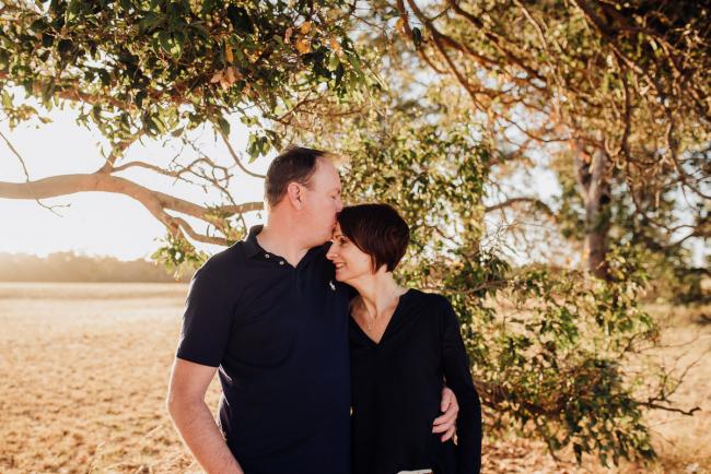 Couple standing arm in arm under a tree with the man kissing his wife's head during an extended family photography session at Perry's Paddock in Perth