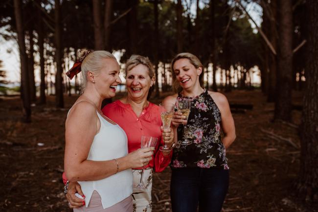 Three women standing with champagne glasses and laughing during an extended family photography session at The Pines Wanneroo in Perth