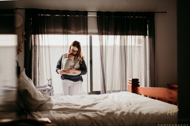 20180610-DSC_9029 mother stands by the window with her new baby sleeping in a baby carrier during Perth newborn photography session