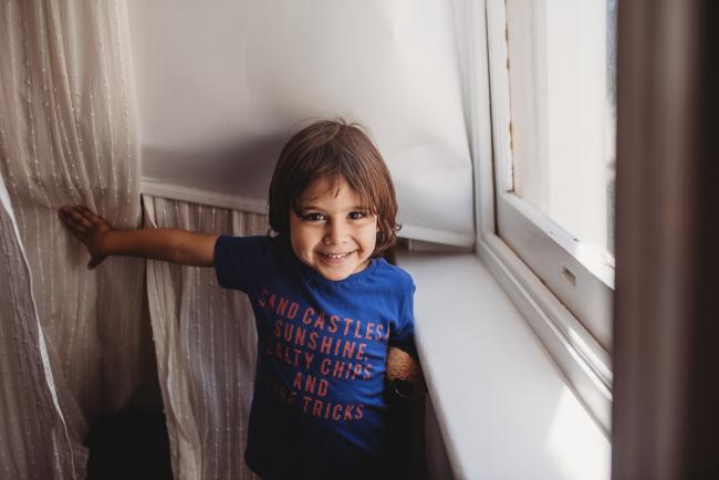 Little boy smiling by the window as he pulls open the curtains with Perth family photographer
