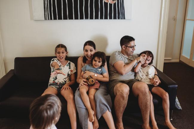 Family of 5 sitting on the couch, with the daughter reaching towards their younger brother who is standing and looking at them with Perth family photographer