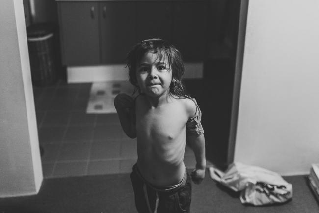 Black and white image of little boy looking at the camera as he pulls his top off with Perth family photographer