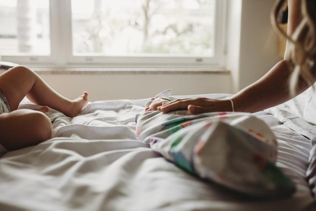 New baby lays wrapped on the bed with mothers hand on her tummy and big sisters feet in the frame during a Perth Fresh 48 photography session