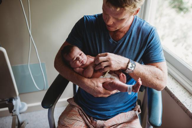 Dad holding his new baby on a hospital chair and touching her hands during Fresh 48 Photography Perth session