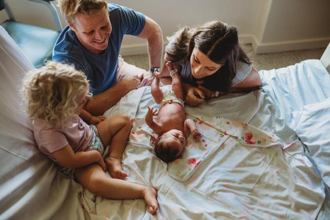 New baby lays on the hospital bed with big sister next to her and mother and father looking over her during a Perth Fresh 48 photography session