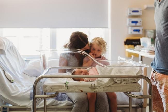 Mother sits with little girl on her lap while looking at the bassinet with their new baby in it during a Perth Fresh 48 photography session