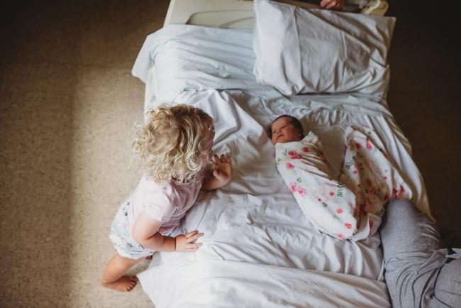 Top down image of big sister leaning against the bed and looking at her baby sister who is wrapped on the hospital bed during Fresh 48 Photography Perth session