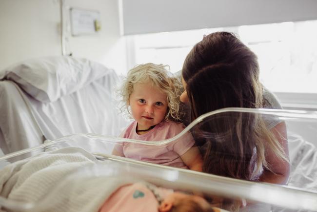 Little girl looks at the camera over her baby sister's hospital bassinet during a Perth Fresh 48 photography session