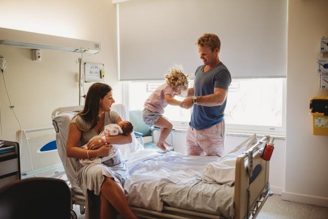 Mother holding new baby on the hospital bed while big sister holds hands with her father and jumps on the bed during Fresh 48 Photography Perth session
