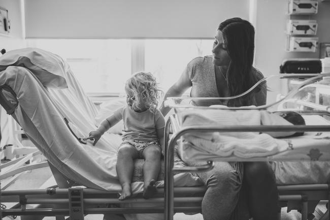 Black and white image of little girl sitting next to her mother and baby sister on a hospital bed during Fresh 48 Photography Perth session