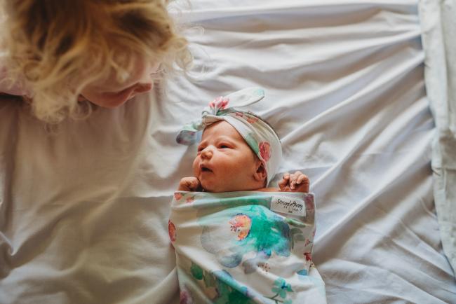 New baby in a floral wrap on a white bed with her big sisters head peeking in the corner to look at her during Fresh 48 Photography Perth session