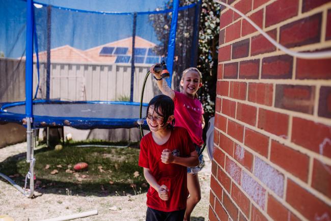 Boy in red top running away laughing from girl who is spraying him with water with Perth family photographer