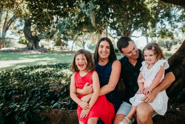 Family-Photographer-Perth-Hyde-Park-1-of-34 Mother and father sitting on a tree with their daughters on their knee during a Perth family photography session at Hyde Park