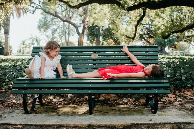 Family-Photographer-Perth-Hyde-Park-11-of-34 Two young sisters on a green park bench - one laying down and one sitting and smiling at her during a Perth family photography session at Hyde Park