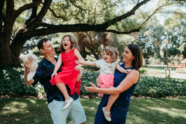Family-Photographer-Perth-Hyde-Park-31-of-34 Mother and father holding their two daughters as they all laugh and reach for a bunny during a Perth family photography session at Hyde Park