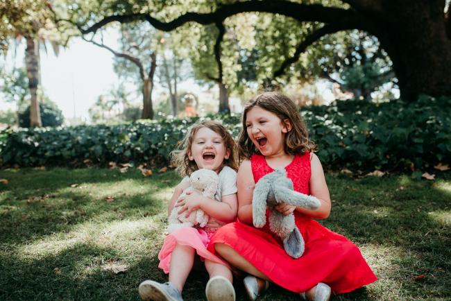 Family-Photographer-Perth-Hyde-Park-32-of-34 Two little girls laughing as they sit on the grass and hold their bunnies during a Perth family photography session at Hyde Park